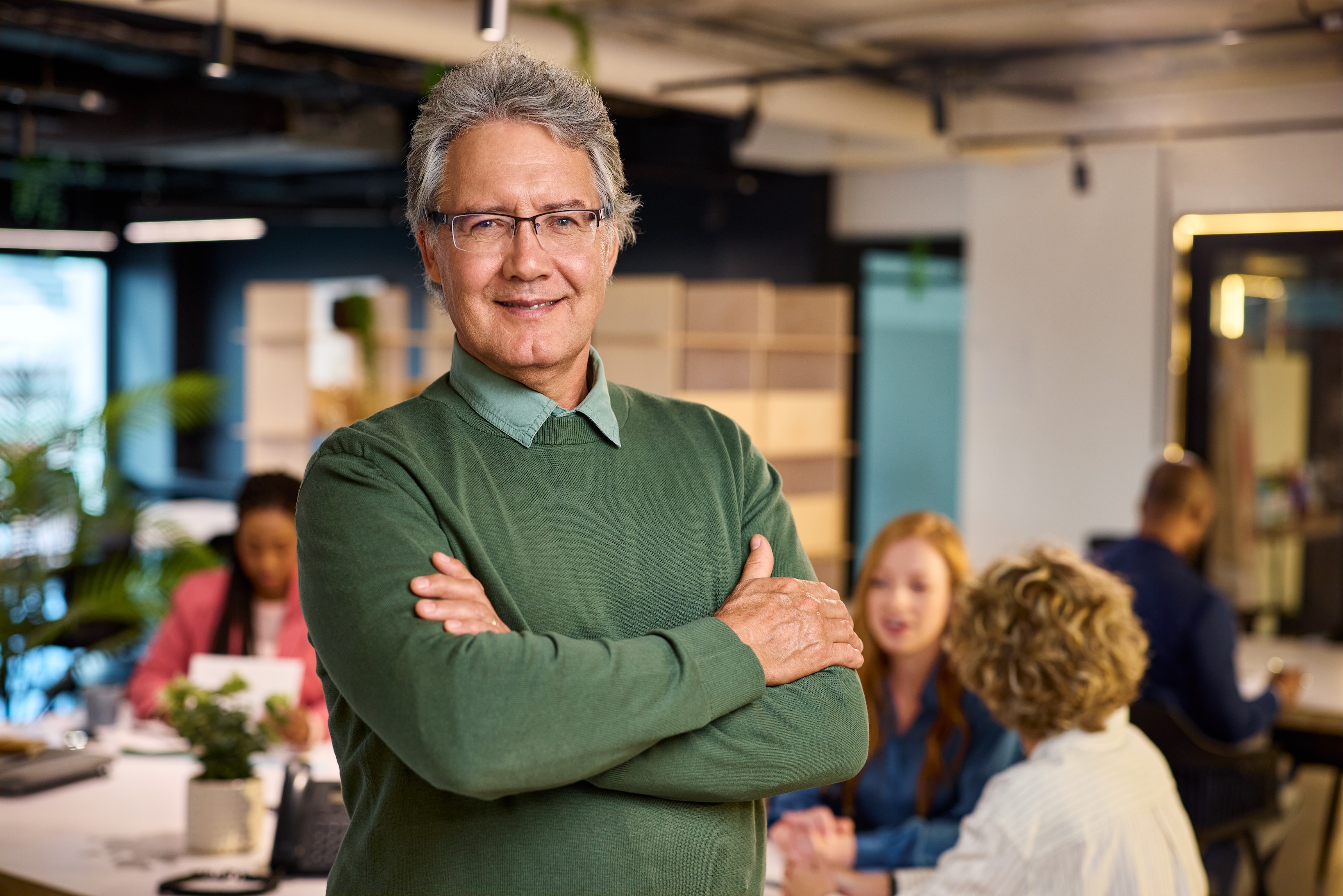 Portrait of reliable mature business man standing in modern office while his teamwork working in background. Successful mature entrepreneur in casual outfit looking at camera with satisfaction. Confident old man with eyeglasses standing with crossed arms with copy space.